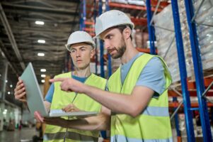 Decision. Two serious focused men in protective helmet and workwear working looking at laptop together pointing at screen at warehouse