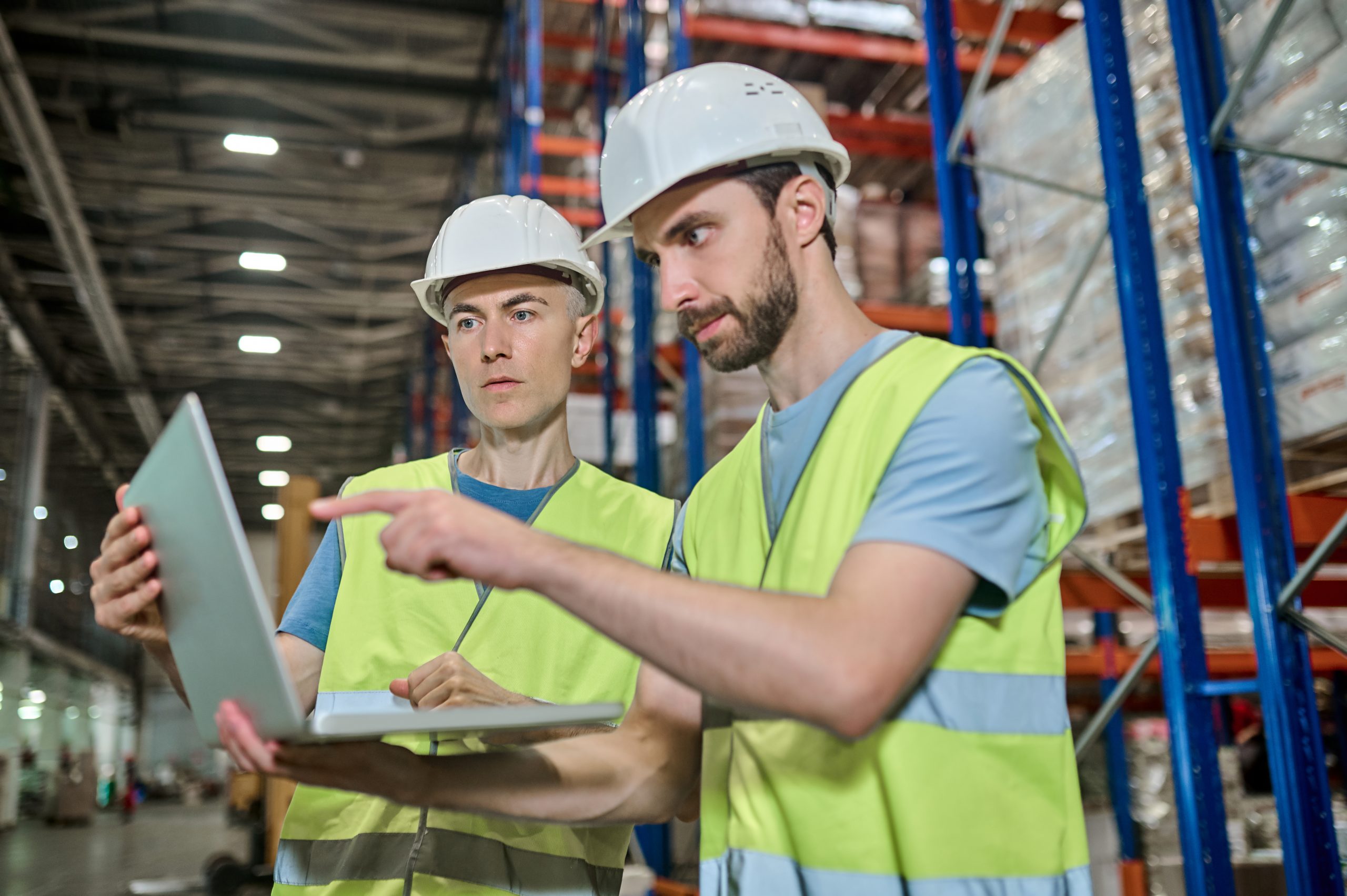 Decision. Two serious focused men in protective helmet and workwear working looking at laptop together pointing at screen at warehouse
